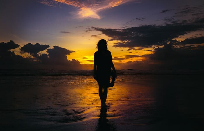 夕日に向かって海辺を１人歩く女性の後ろ姿の哀愁漂うシルエット